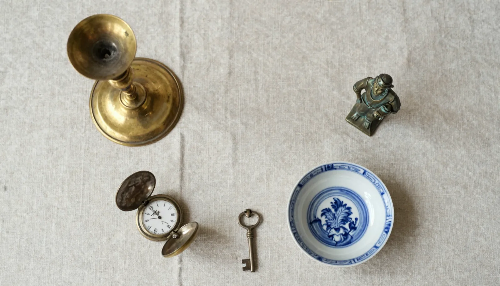 Flat lay of assorted antiques: brass candlestick, bronze figurine, open pocket watch, transferware bowl and an old key