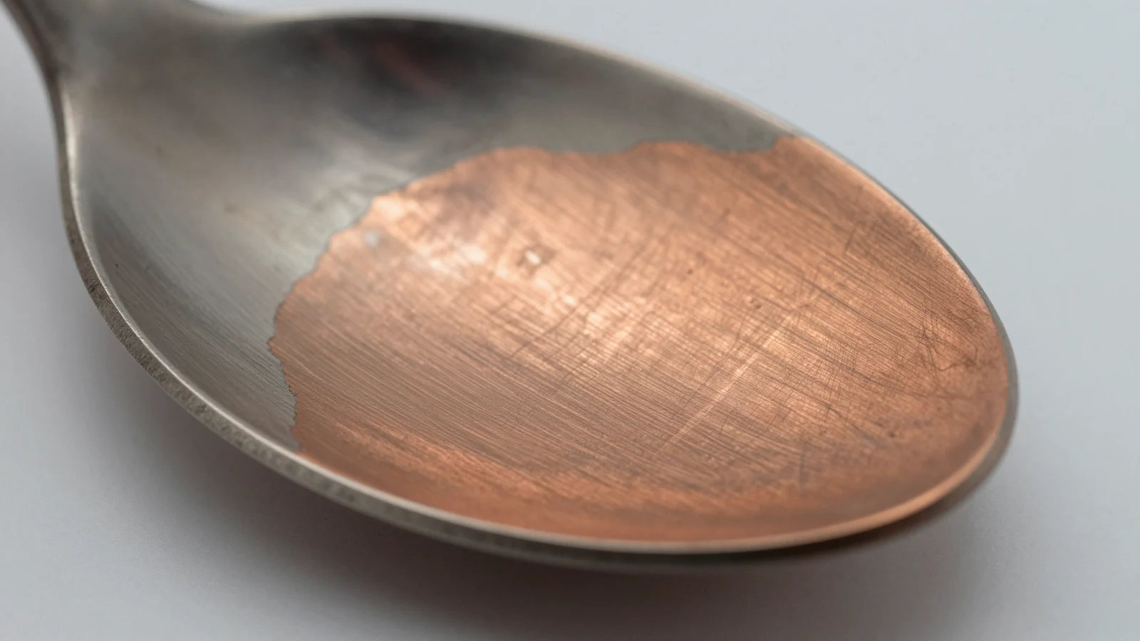 Close-up of a spoon bowl where the silvery surface has worn away across one side to a reddish copper colour underneath